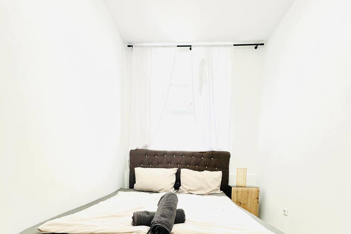 A single bed with a dark headboard and footboard, a wooden nightstand, and a window with sheer curtains.