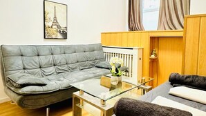 Een moderne woonkamer met een grijze bank, een glazen salontafel, een bed met donkergrijze beddengoed en een houten kast.
