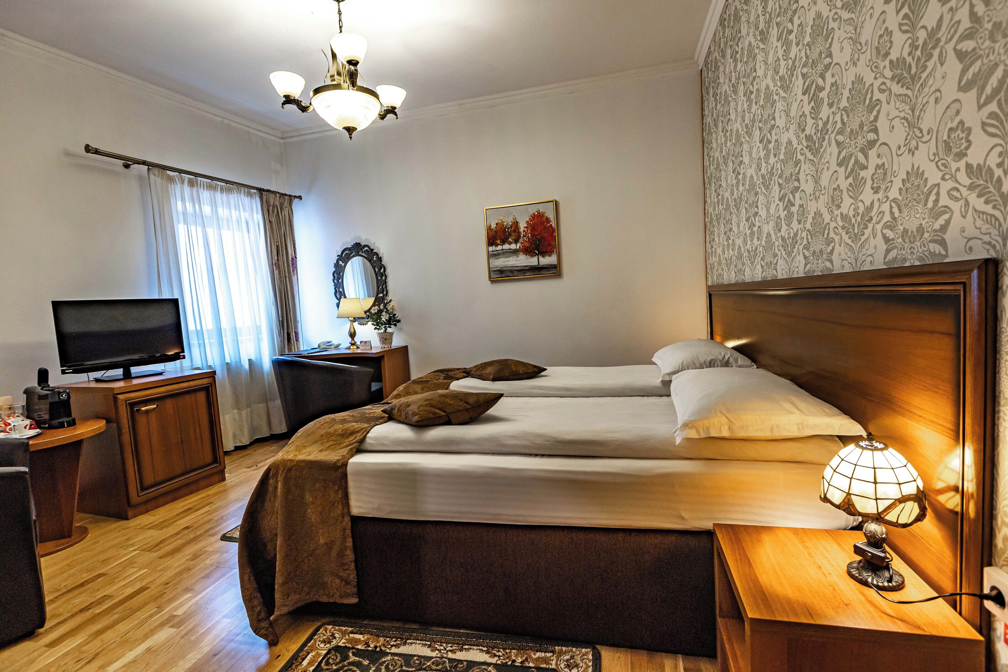 A hotel room with a bed, bedside table, lamp, TV, desk, and a window with curtains.
