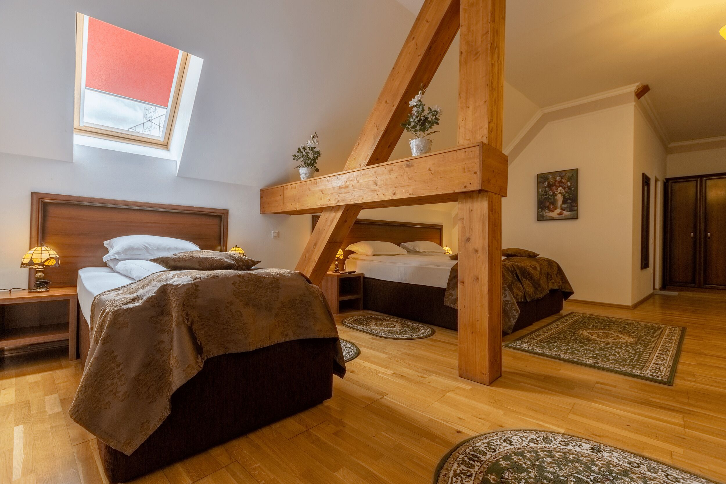 Une chambre avec un plafond en pente, des poutres en bois, un puits de lumière, deux lits, des tables de chevet, des lampes et un tableau au mur.