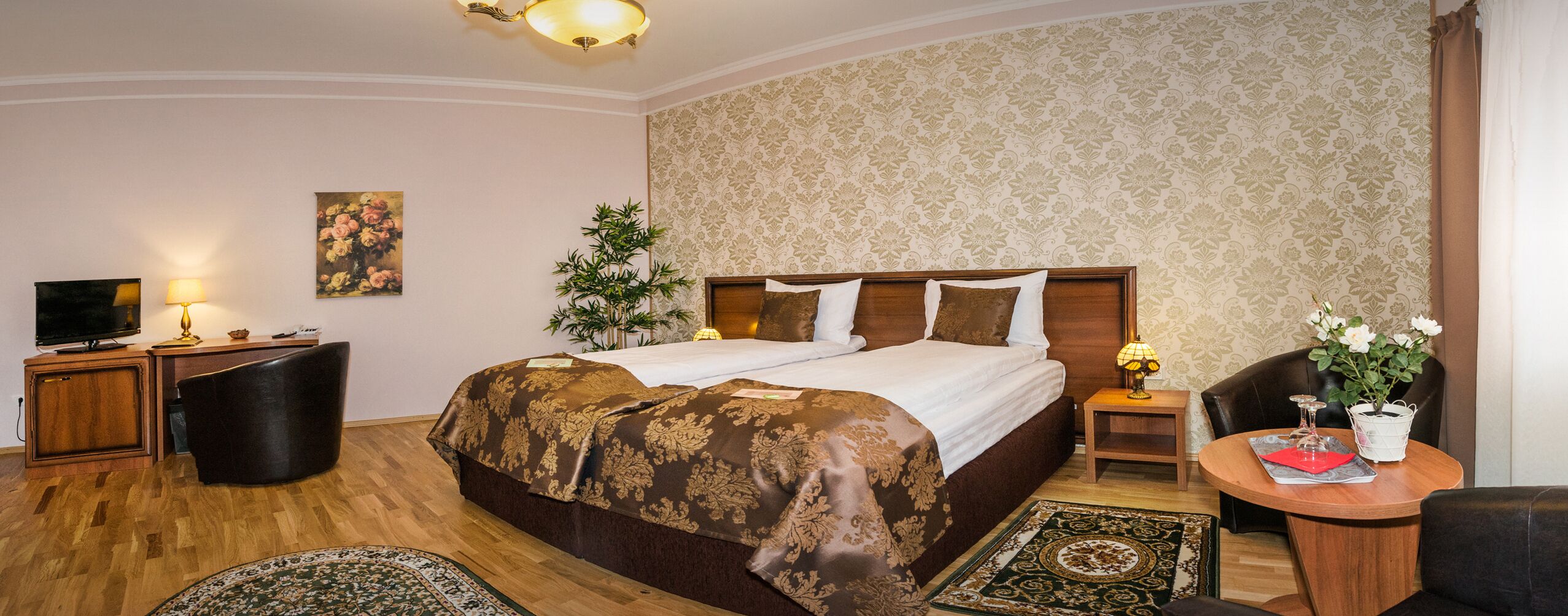 A hotel room with a large bed, a television, a desk with a lamp, and a small table with a vase of flowers.