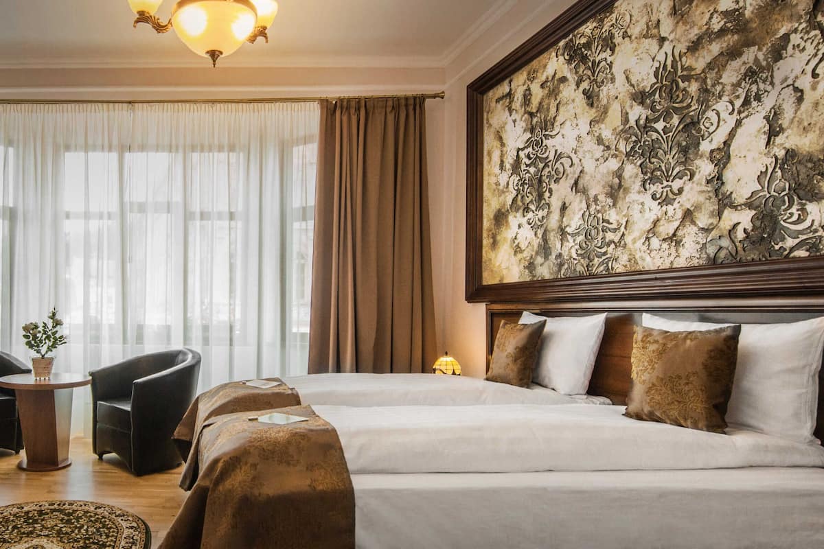 Habitación de hotel con una cama grande, dos sillones, un escritorio con lámpara y un ventanal con cortinas.