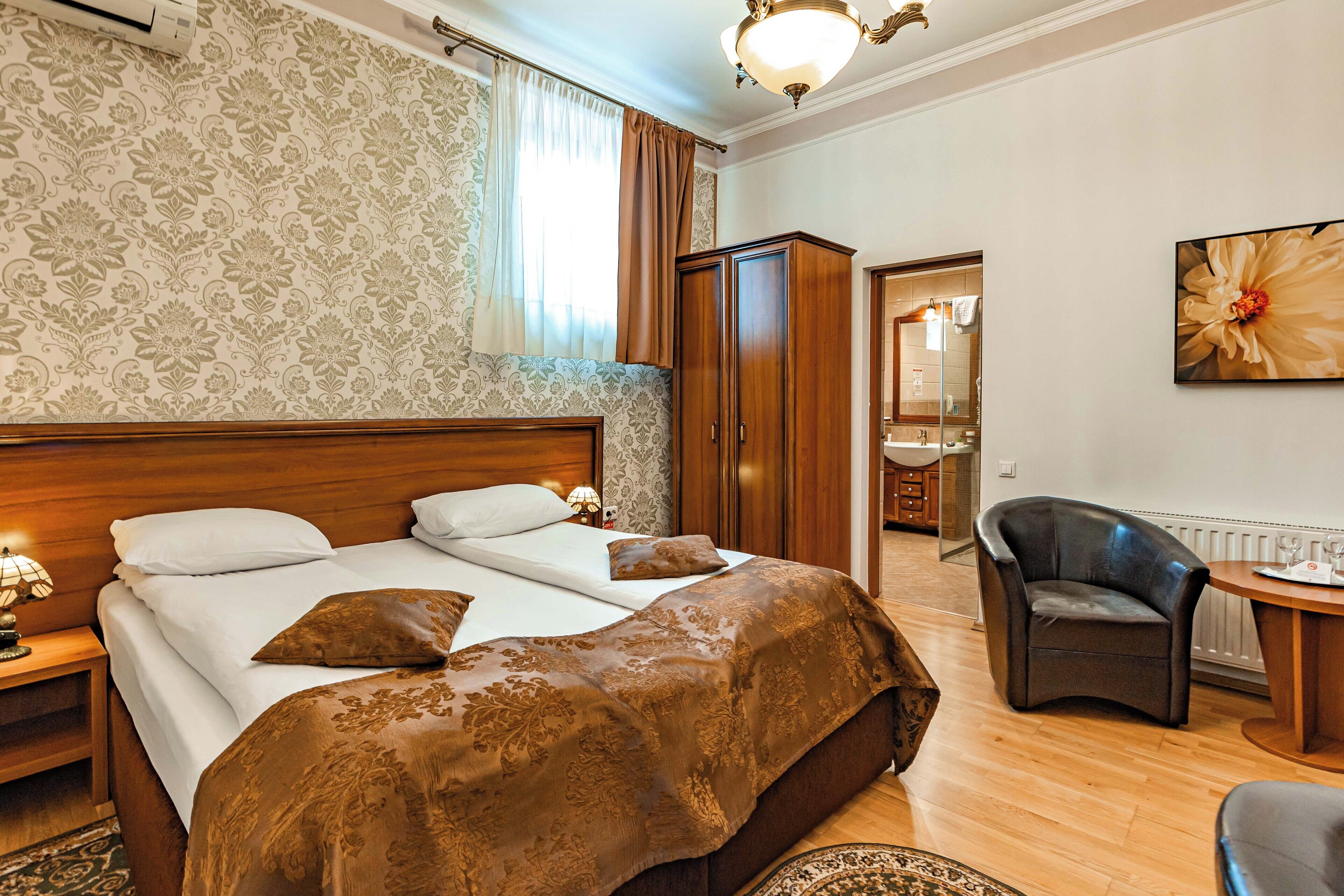 A hotel room with a large bed, a wooden wardrobe, a desk with a chair, and a large window with curtains.