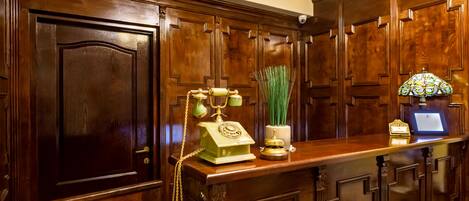 Un área de recepción con un mostrador de madera, un teléfono antiguo, un jarrón con plantas verdes y una lámpara decorativa.