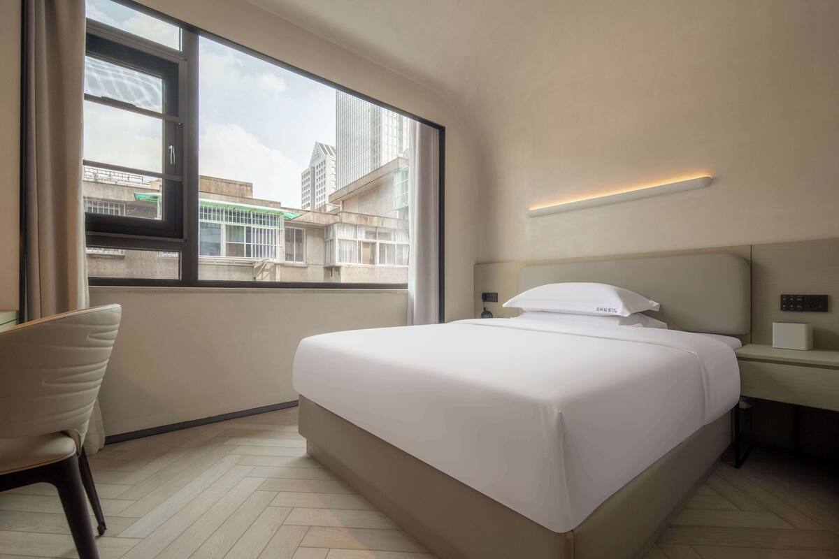 A hotel room with a large window, a bed, a chair, and a bedside table with a lamp.