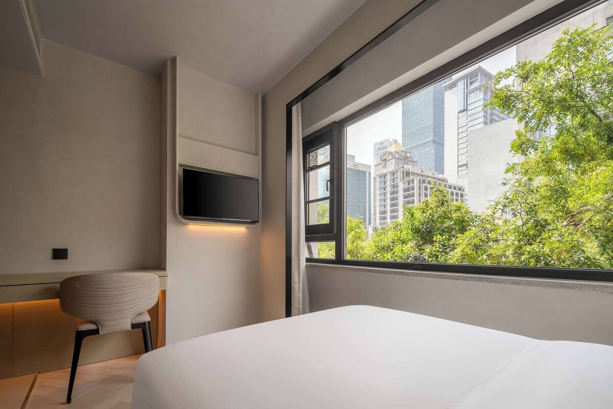 A modern hotel room with a large window, a bed, a chair, and a flat-screen TV mounted on the wall.