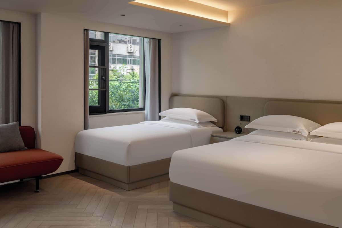 A hotel room with two beds, a red sofa, a window with a view of greenery, and a modern ceiling light fixture.
