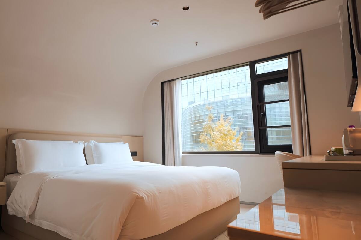 A hotel room with a bed, a nightstand, a window with blinds, and a view of a building.