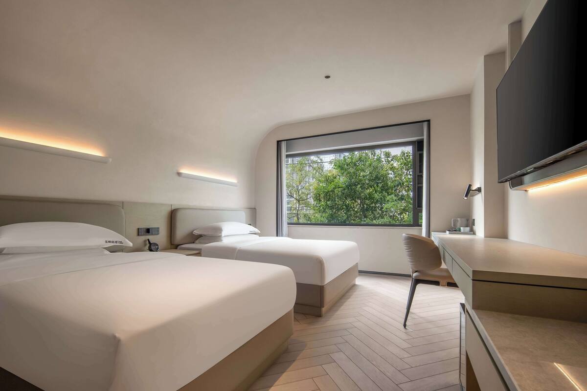 A modern hotel room with two beds, a desk, and a large window offering a view of greenery.