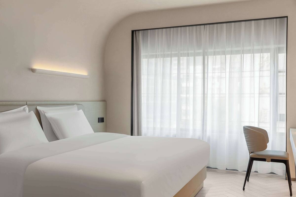 A modern hotel room with a large bed, a chair, and a large window with sheer curtains.
