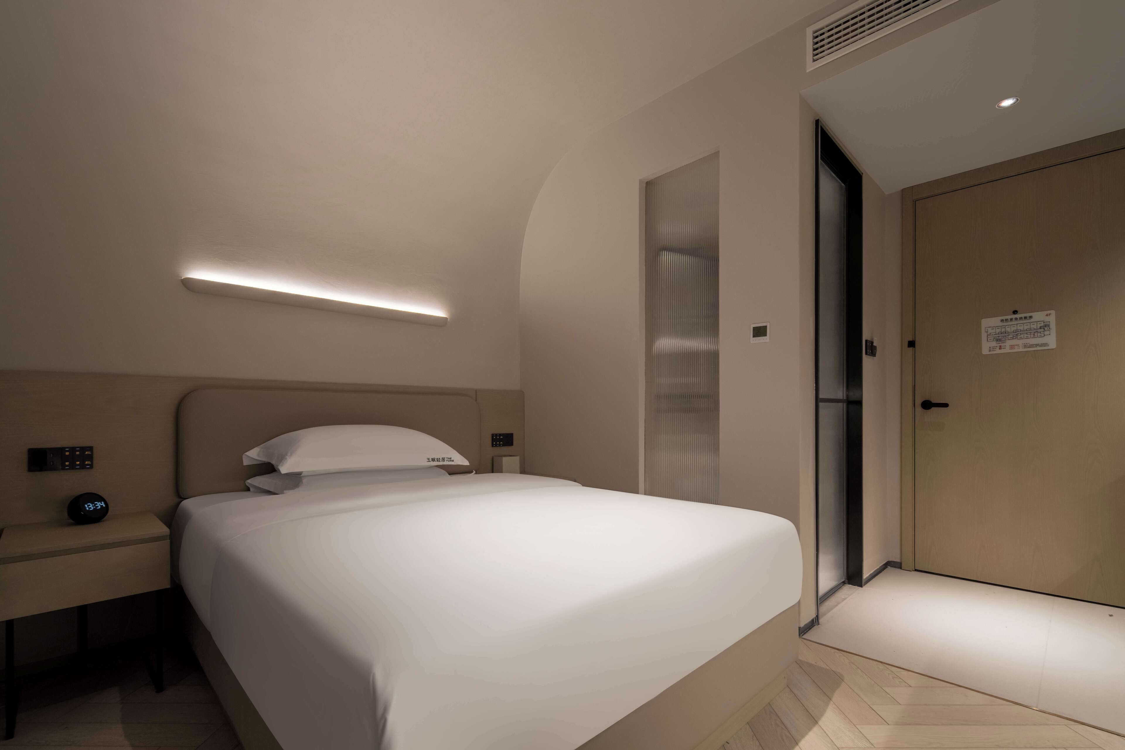 A modern hotel room with a neatly made bed, a bedside table with a clock, and a door with a sign.