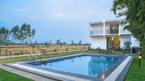 A rectangular swimming pool with a surrounding deck, a two-story building with balconies, and a grassy area with trees in the background. - Evencia Resort (Ramnagar)