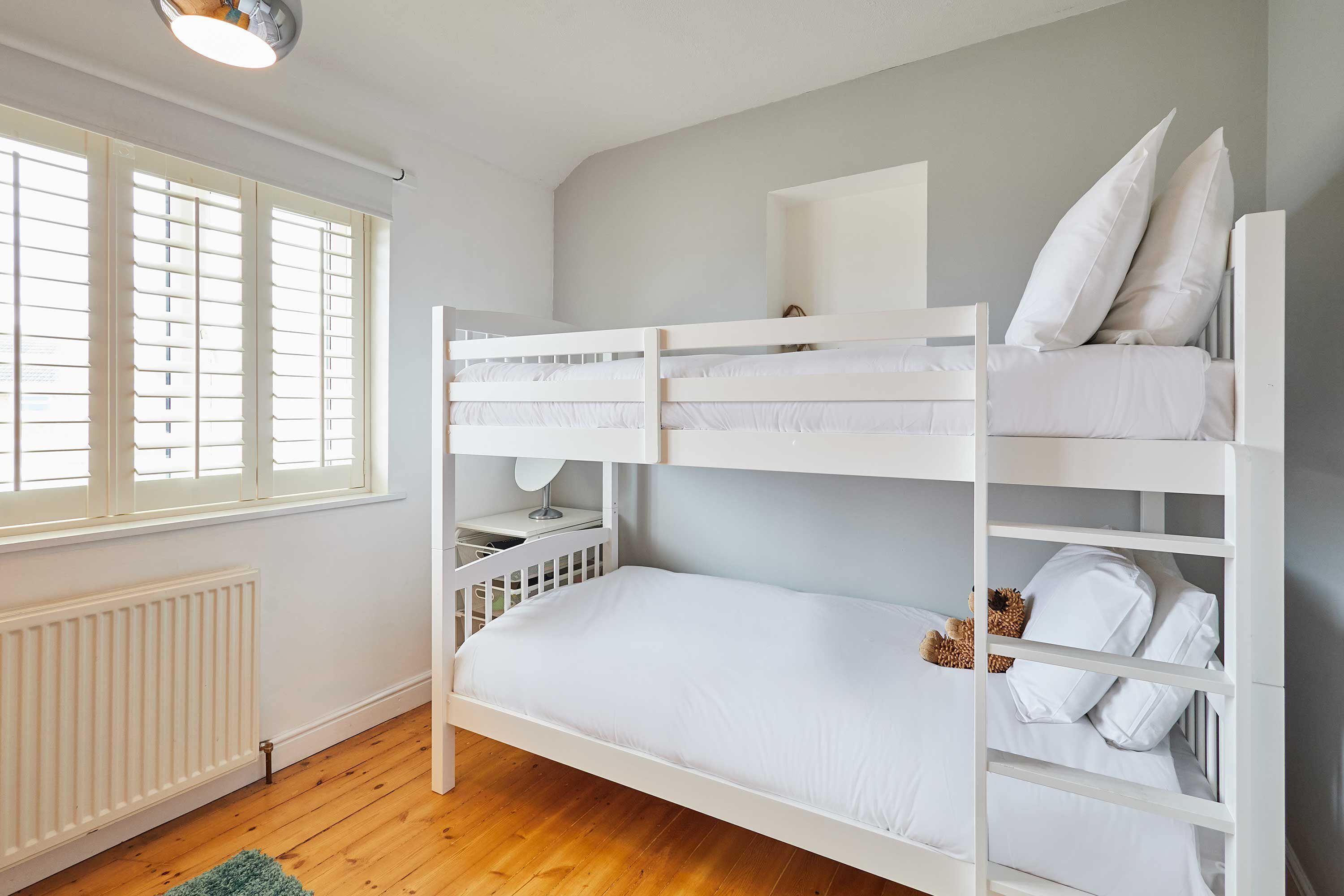 Een slaapkamer met stapelbedden, witte beddengoed, een tafeltje en een raam met jaloezieën.