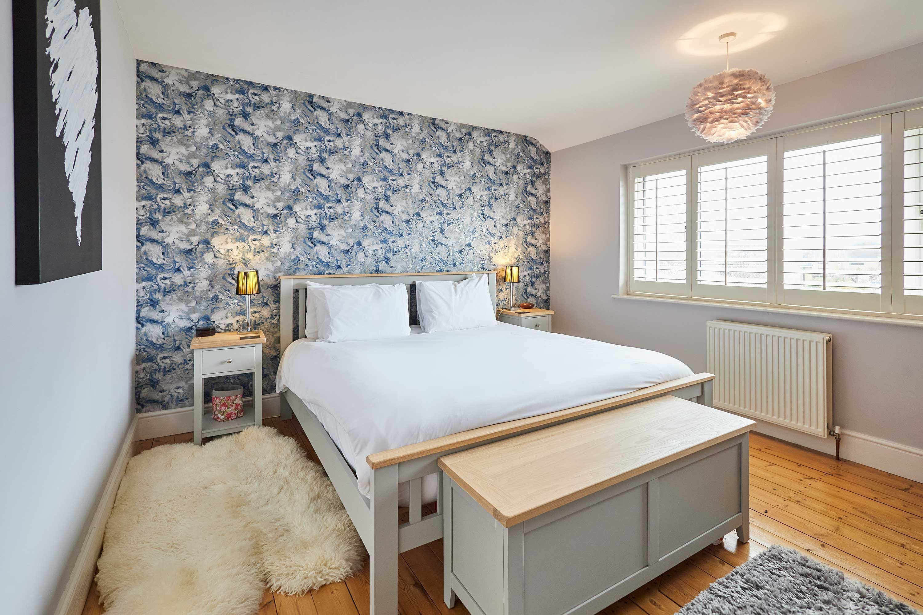 A bedroom with a bed, bedside tables, a bench, a fluffy rug, a radiator, and a window with blinds.