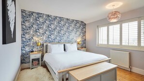 A bedroom with a bed, bedside tables, a bench, a fluffy rug, a radiator, and a window with blinds. - Host Stay Chestnut Close (Saltburn-by-the-Sea)