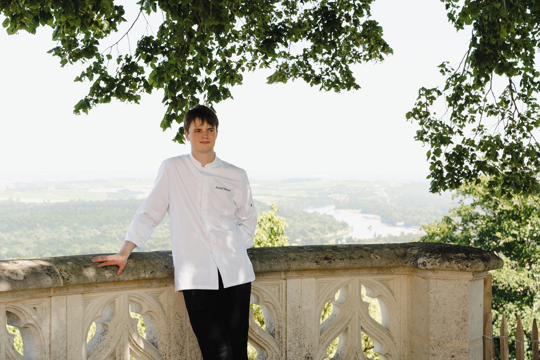 Una persona con bata de chef blanca de pie sobre una barandilla de piedra, con una vista panorámica de un valle y un río al fondo.