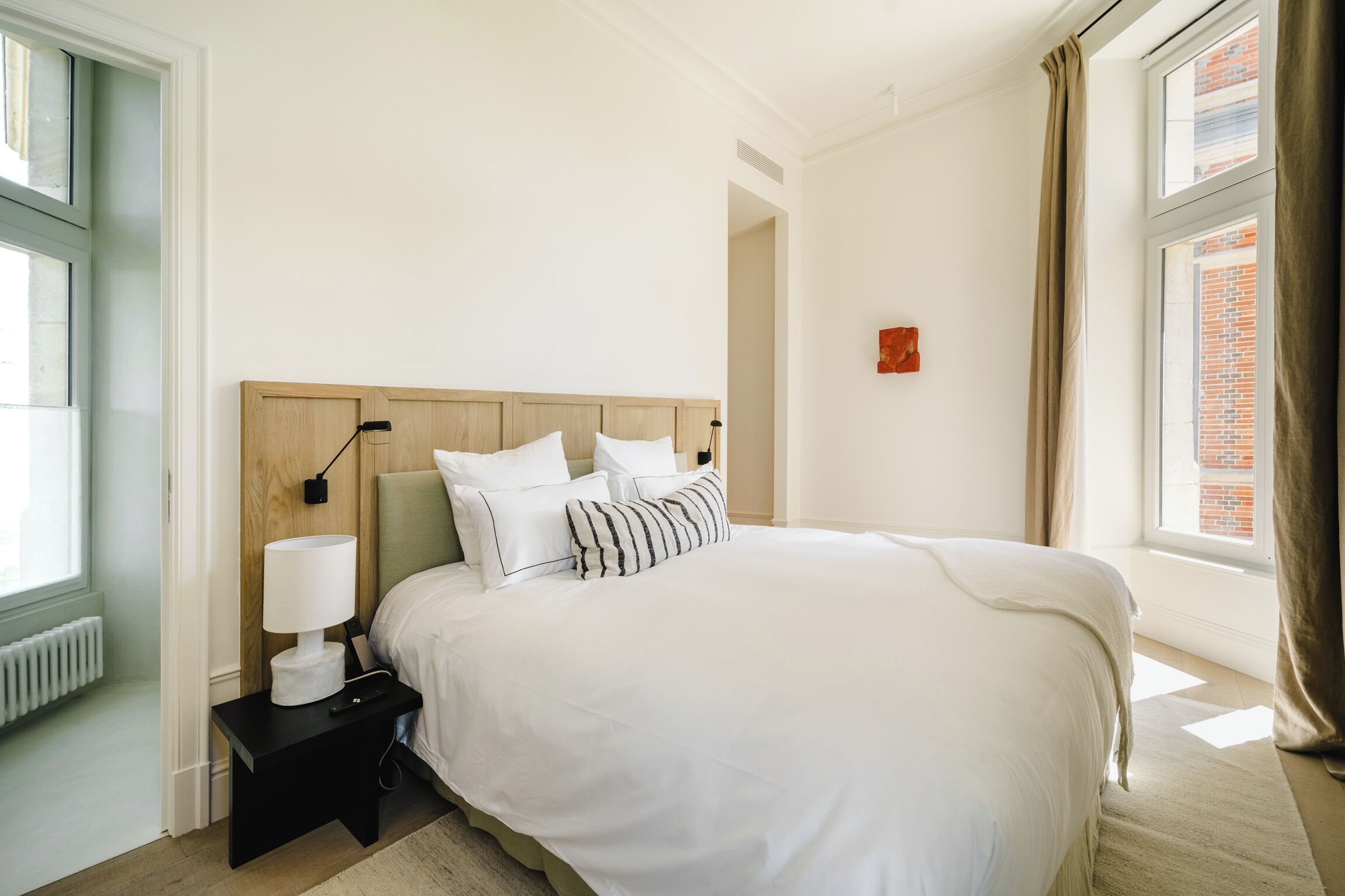A neatly arranged bedroom with a large bed, a bedside table with a lamp, and a view of a brick building through the window.