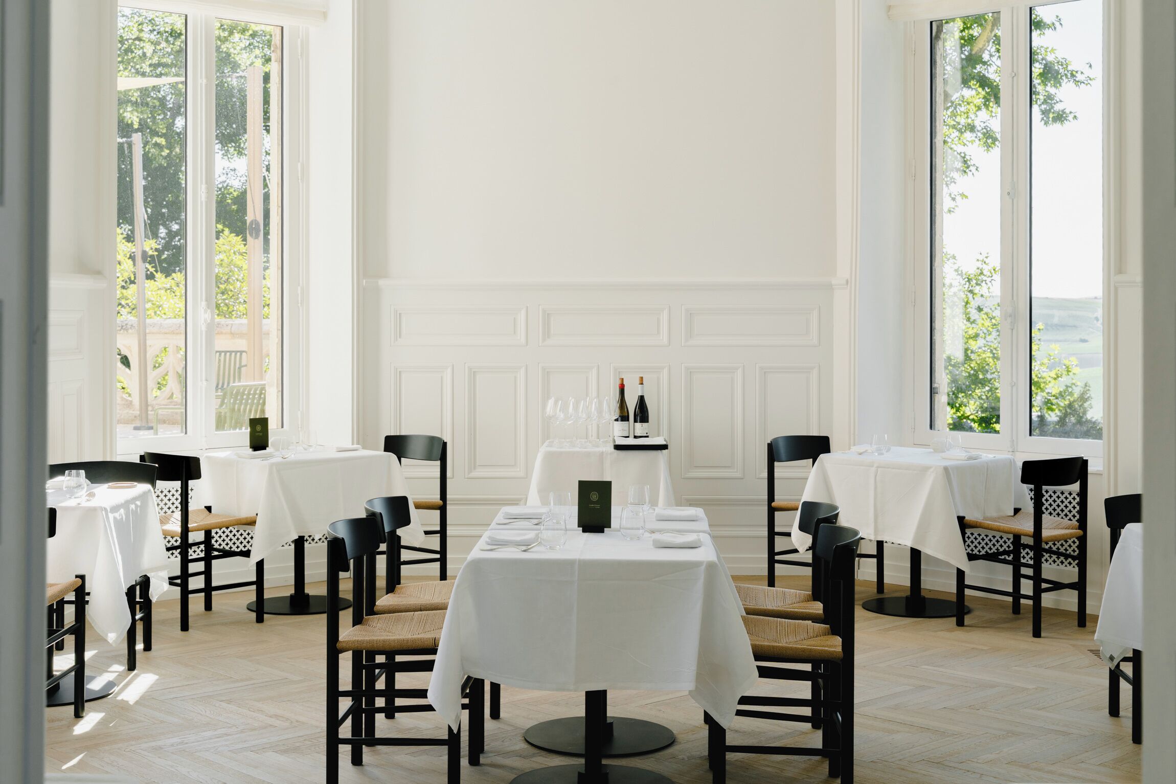 Un comedor con mesas y sillas blancas, un atril para el menú y ventanas grandes con vista a zonas verdes.