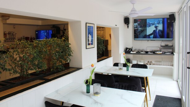 A modern kitchen with a large TV, a dining area with a white table, and a bar area with a plant and a TV screen. - Burghotel - das Original (Nuremberg)