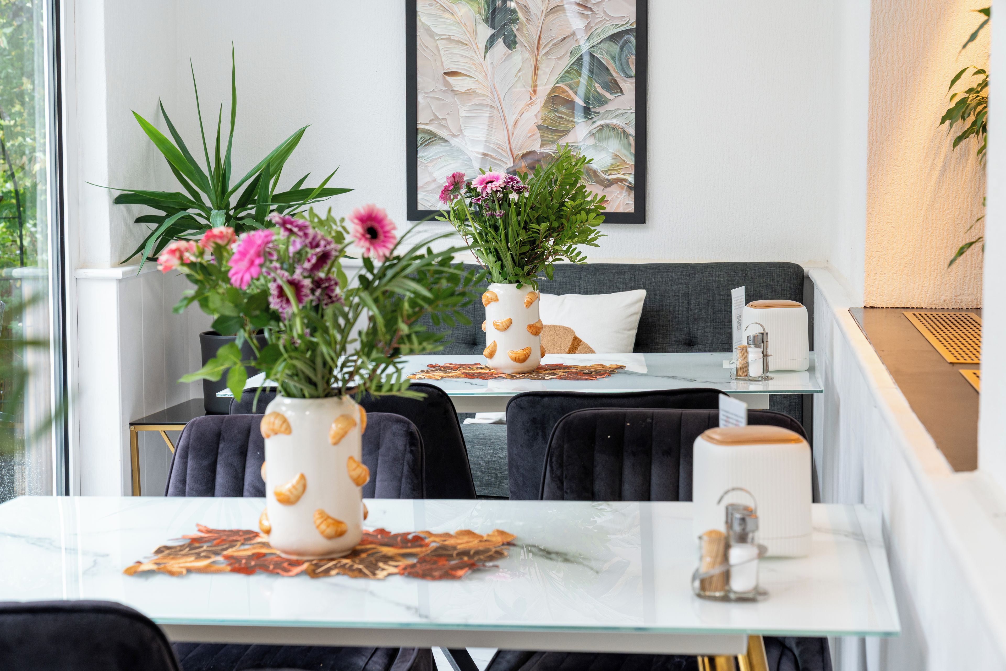 Uma sala de jantar moderna com mesa de vidro, cadeiras pretas e dois vasos com plantas.
