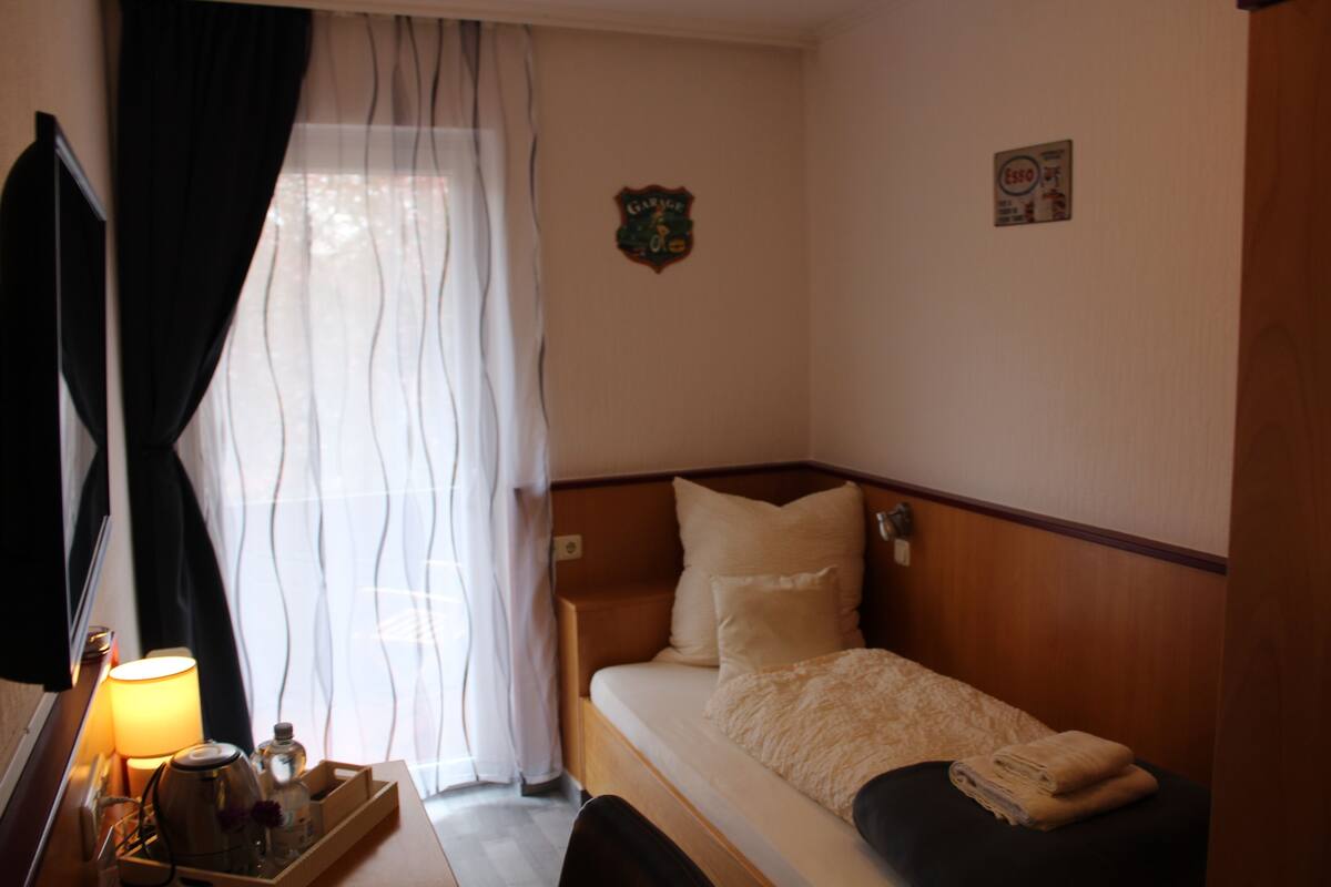 A small, neatly made bed with a wooden headboard, a bedside table with a lamp, and a window with sheer curtains.