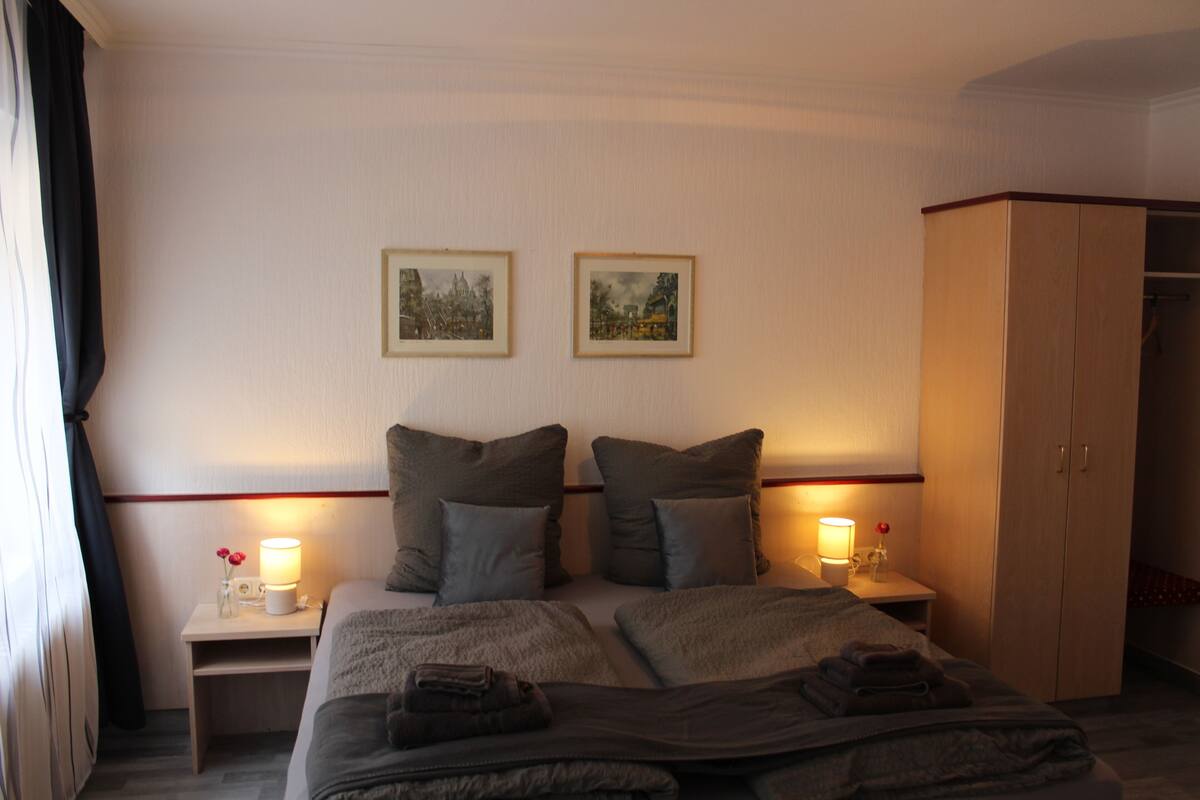 A hotel room with a large bed, two bedside tables with lamps, a wardrobe, and two framed pictures on the wall.