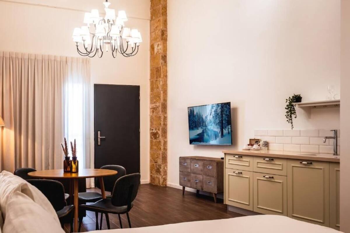 Quarto de hotel com cama, mesa de jantar com cadeiras, lustre, quadro na parede, área de cozinha com armários e uma porta.