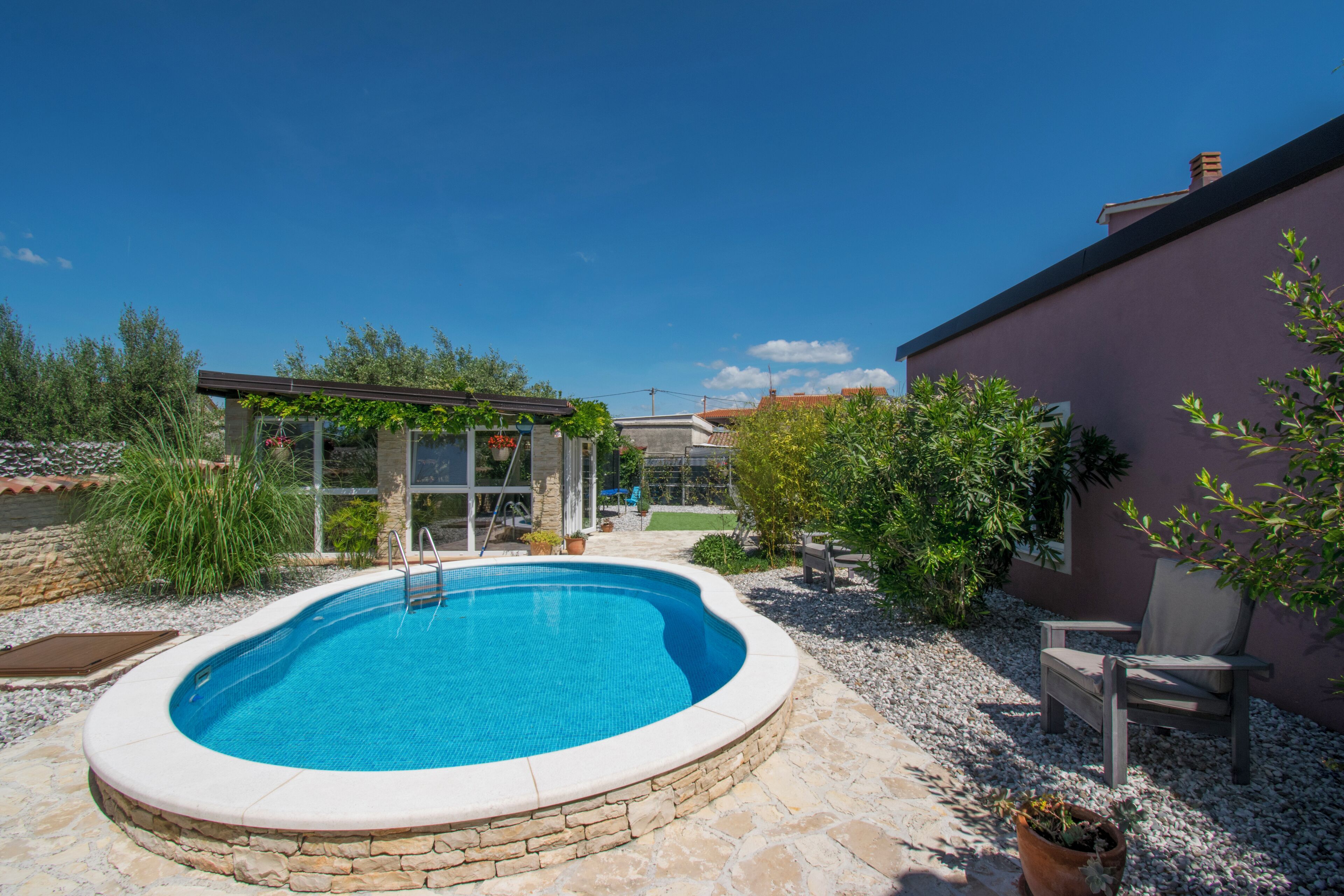 Une piscine entourée d’une terrasse en pierre, une pergola ornée de plantes et un espace patio avec une chaise et une table.