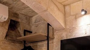A stone-walled room with a bed, a wooden nightstand, a hanging coat rack, and a mounted TV. - Zekka House (Valletta)