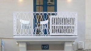 A building with a blue door, a balcony with white railings, and a number '115T' above the door. - Zekka House (Valletta)
