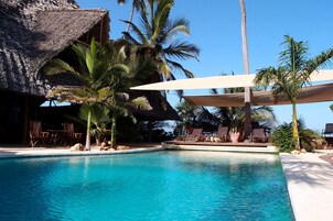 Piscina con sillas de descanso y palmeras, una estructura de techo de paja y un cielo azul despejado.