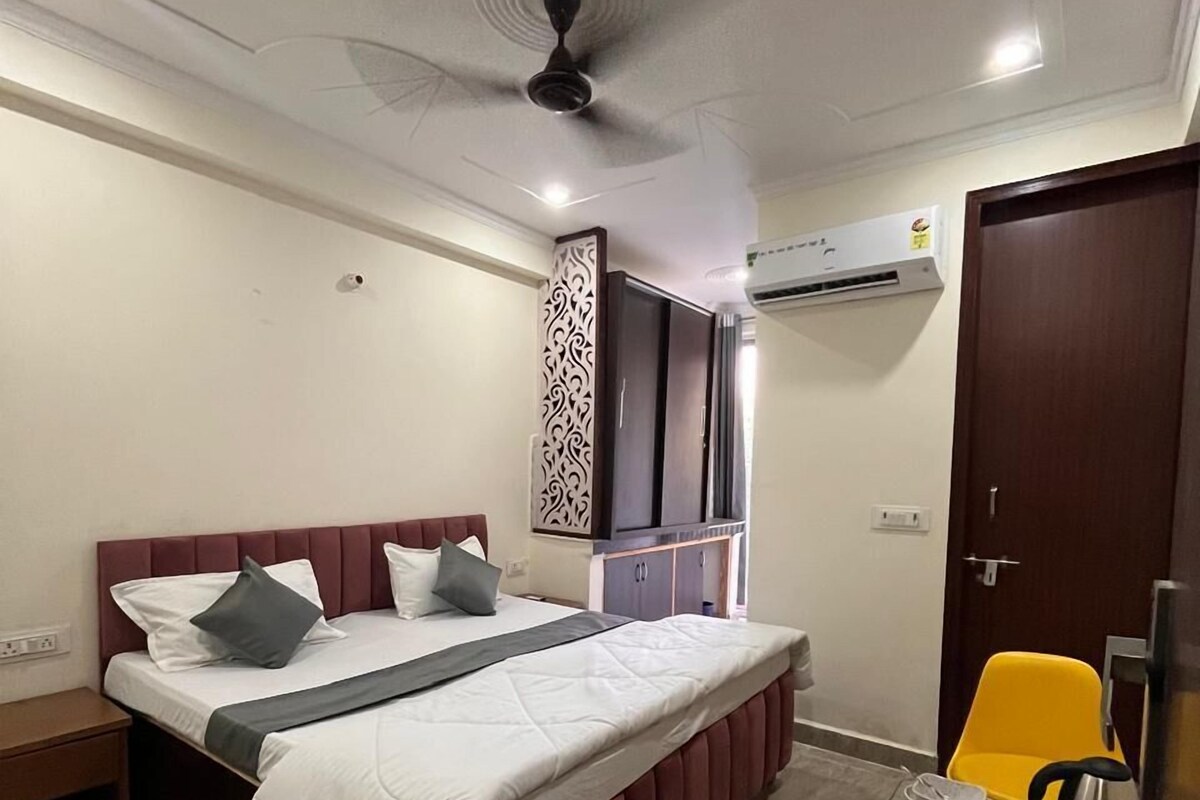 A hotel room with a bed, a desk with a chair, an air conditioning unit, and a door.