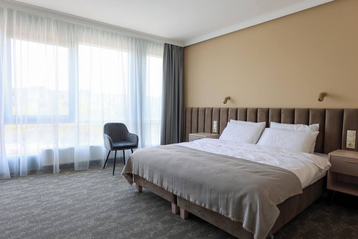 Ein Hotelzimmer mit einem großen Bett, einem Sessel, einem Nachttisch und einem Fenster mit durchsichtigen Vorhängen.