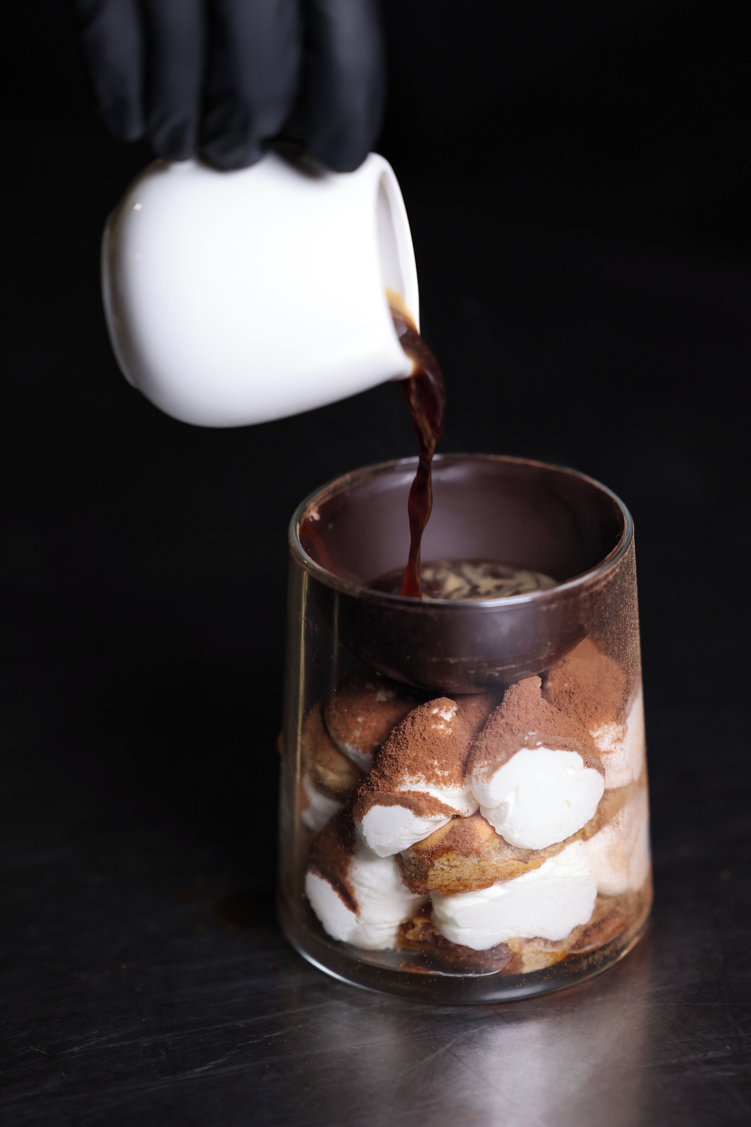 A clear glass mug with a layered dessert, a white cup pouring a dark liquid into it, and a black background.