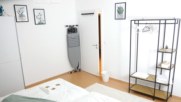 Habitación de hotel con dos camas, un armario, un tendedero y una estantería con toallas y una planta.