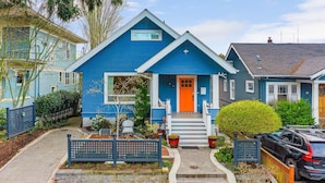 Une maison bleue avec un porche à l’avant, des marches menant à la porte et un petit jardin agrémenté de plantes en pot et d’un banc.