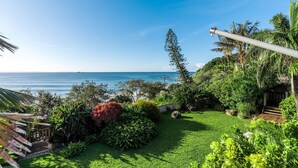Ein üppiger Garten mit einer Holzterrasse, einem hohen Baum und Blick aufs Meer.