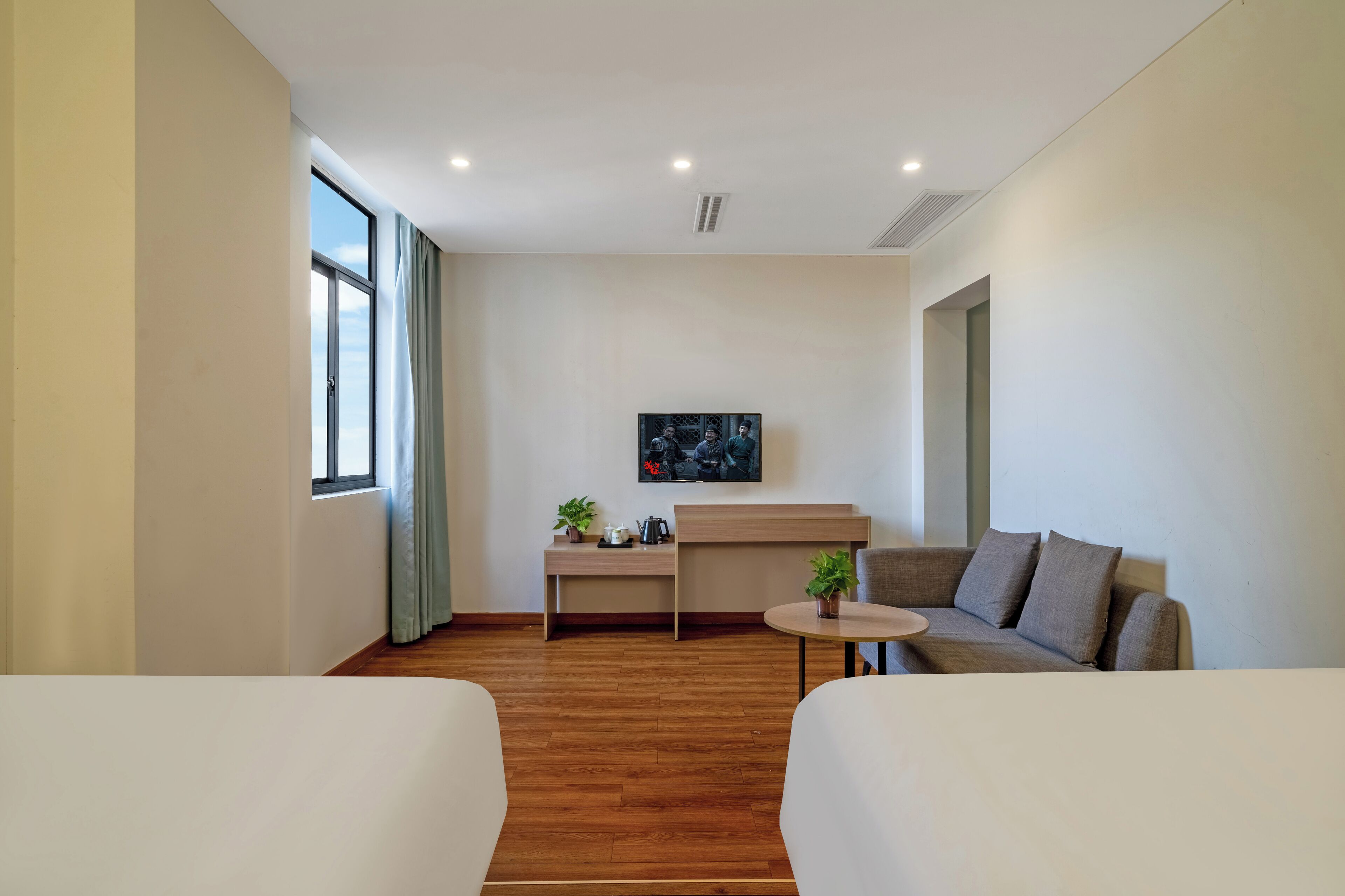 A modern hotel room with a wooden floor, a sofa, a small round table, a desk with a plant, and a painting on the wall.