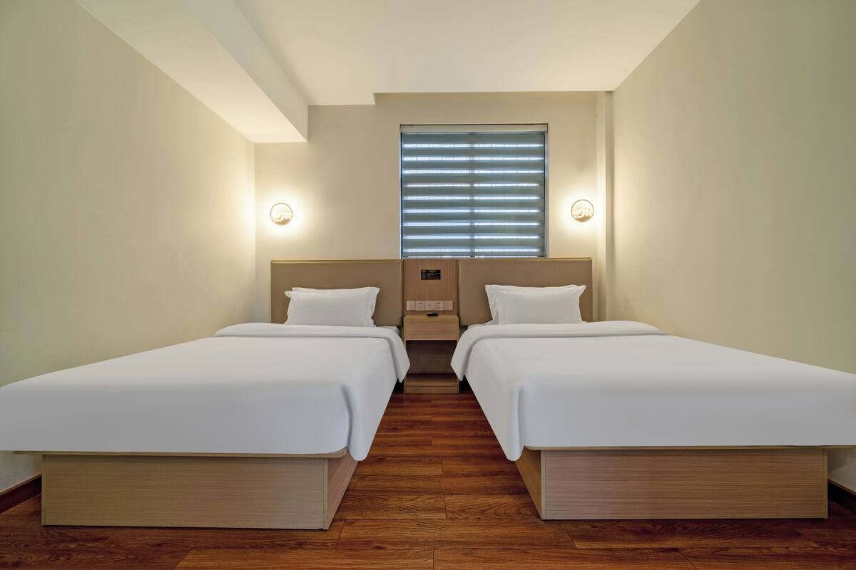 Een hotelkamer met twee enkele bedden, houten vloeren, een groot raam met zonwering en bedlampen.