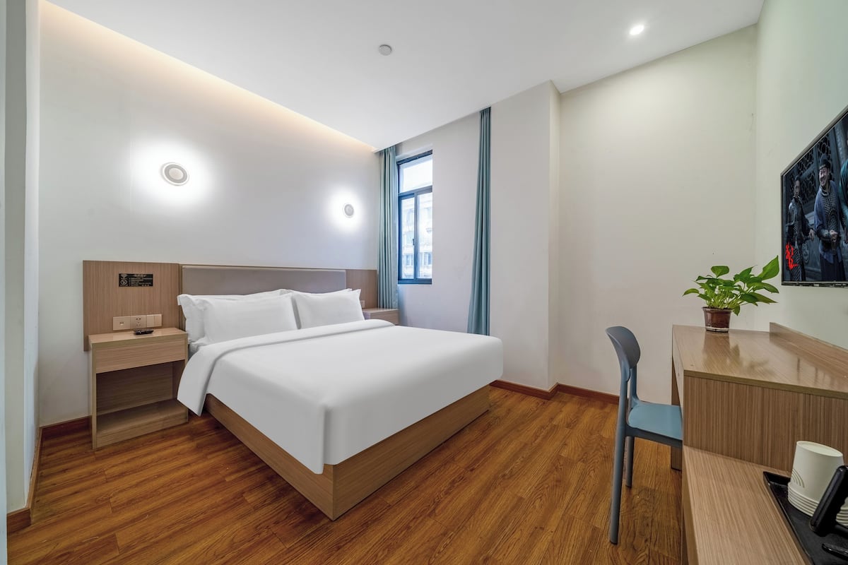 Een moderne hotelkamer met een houten vloer, een groot bed met witte lakens, een bureau met een stoel, en een kamerplant.