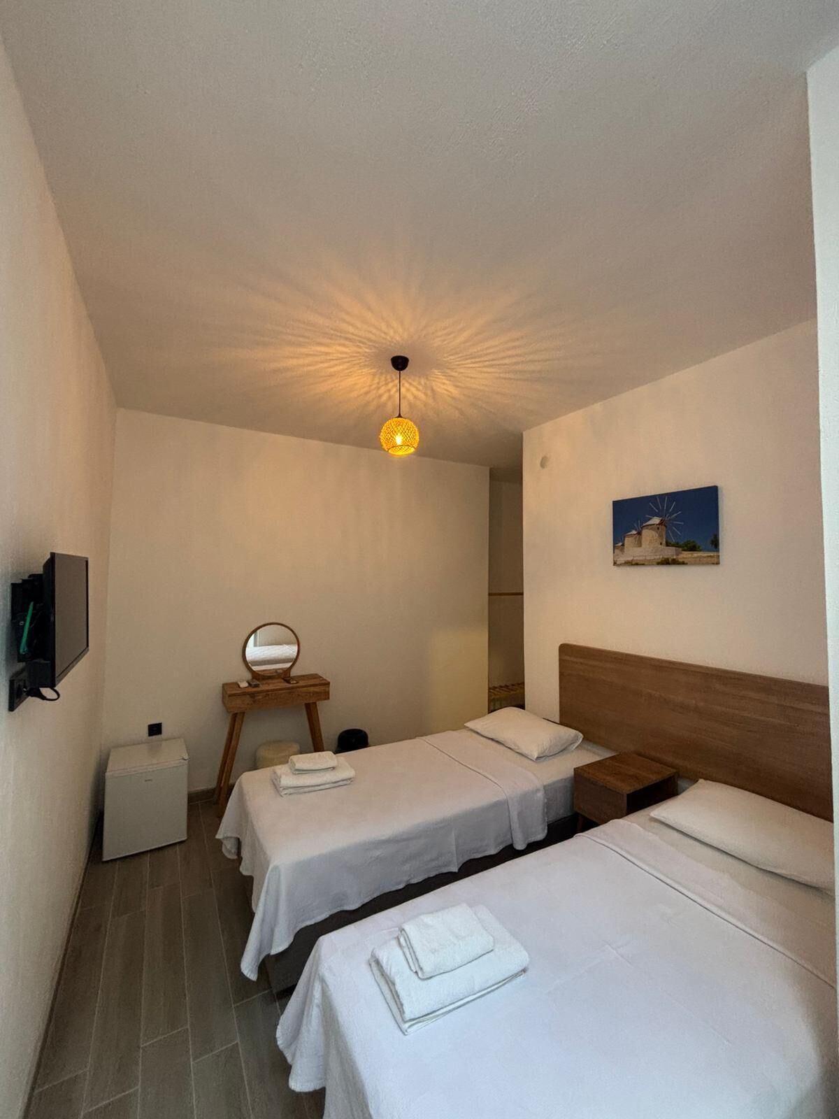 A hotel room with two single beds, a small table, a mirror, a television, and a ceiling light fixture.