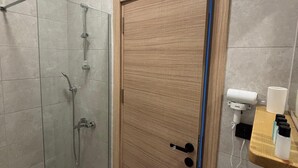 Ein Badezimmer mit Duschkabine aus Glas, einer Holztür, einem weißen Waschbecken und einem Fön auf einem Regal.