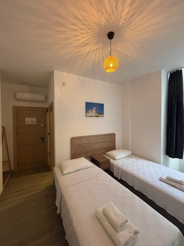 A hotel room with two beds, a wooden headboard, a nightstand, a wall-mounted picture, a window with curtains, and a ceiling light fixture. - Kırçak Konak (Cesme)