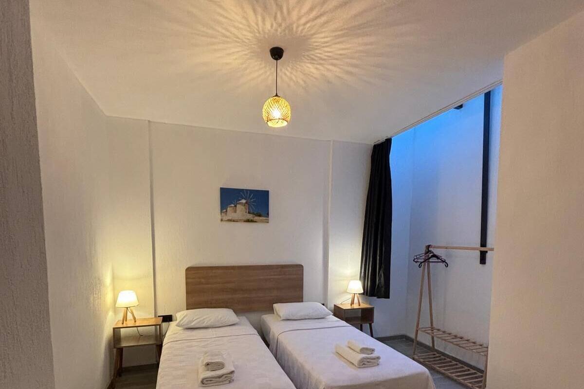 Une chambre d’hôtel avec deux lits, une tête de lit en bois, un luminaire au plafond, une petite table et une fenêtre avec des rideaux.
