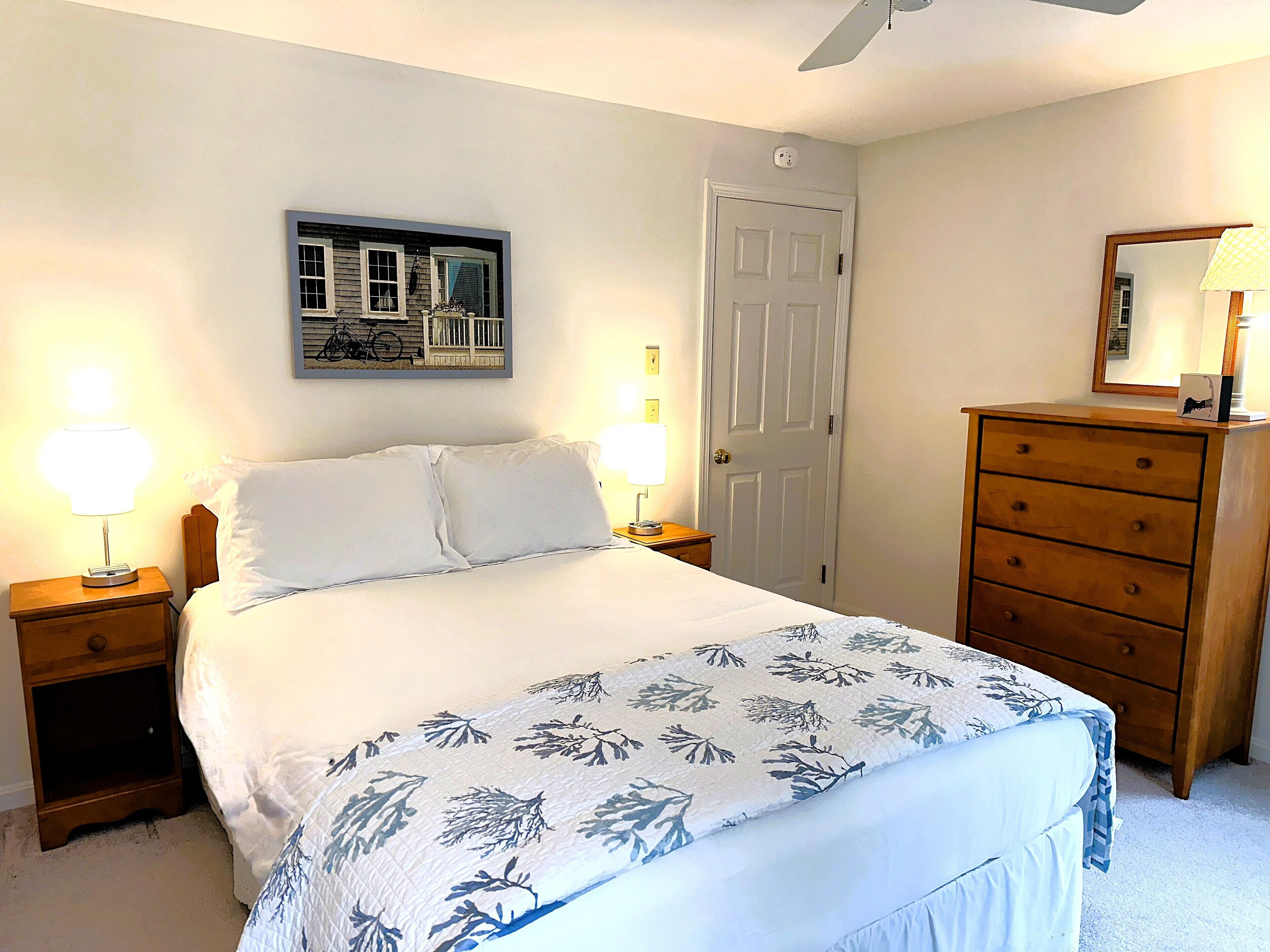 A neatly made bed with a floral bedspread, two bedside lamps, a wooden dresser with a mirror, and a door with a light switch above it.