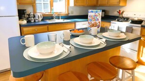 Une cuisine avec des meubles de rangement en bois, un plan de travail bleu et un coin repas comprenant des bols, des tasses et une boîte de céréales.