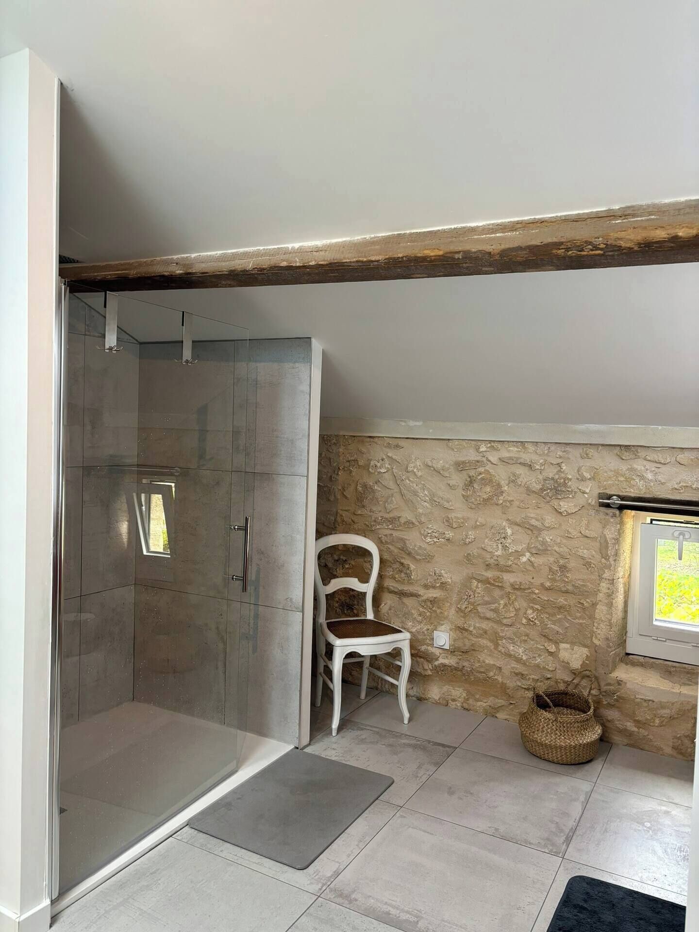 A bathroom with a glass shower enclosure, a white chair, a stone wall, and a window with a view.