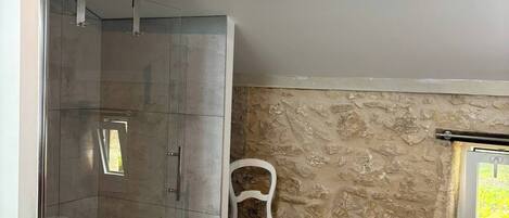 A bathroom with a glass shower enclosure, a white chair, a stone wall, and a window with a view.