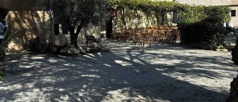 A courtyard with a gravel ground, a pergola with chairs, and a building with a tiled roof.