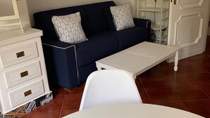 Un salon avec un canapé bleu foncé, une table basse blanche et un meuble blanc. On aperçoit un panier de fruits sur une table blanche au premier plan.